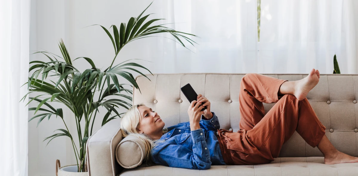Blonde Frau in Jeanshemd liegt auf einem beigen Sofa und lächelt in ihr Smartphone