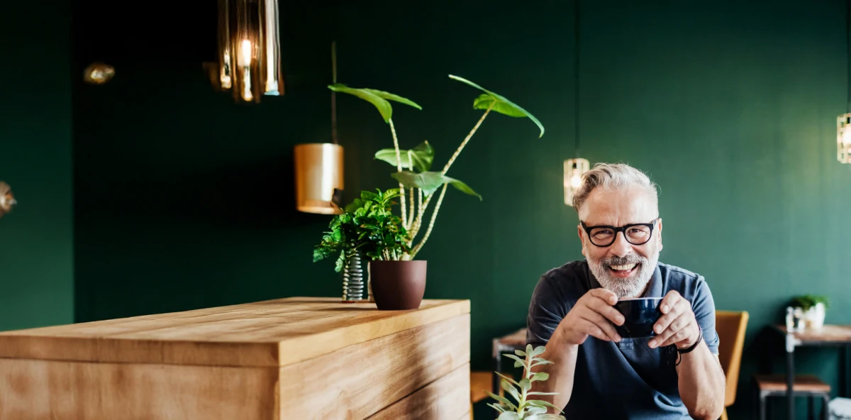 Grauhaariger Mann mit schwarzer Brille sitzt vor einer Grünen Wand mit einer Tasse in der Hand
