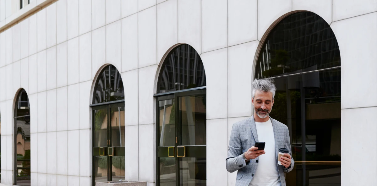 Mann mit grauen Haare und Bart in Sakko steht mit Kaffee to Go und Smartphone in der Hand vor einem Gebäude