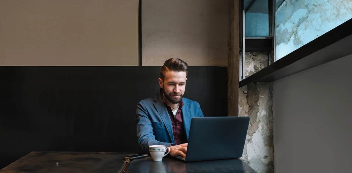 Braunhaariger Mann in blauem Sakko sitzt an seinem Laptop neben einer Tasse Kaffee