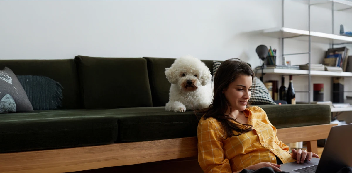 Braunhaarige Frau sitzt vor Sofa auf dem Boden und arbeitet am Laptop während kleiner weißer Hund hinter ihr auf dem Sofa liegt