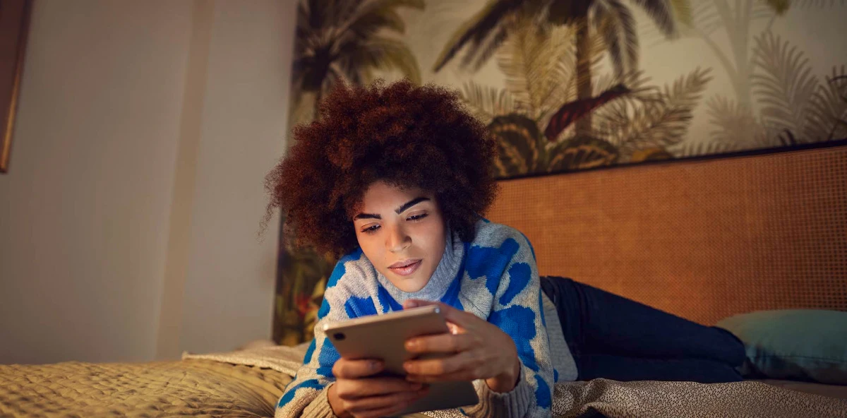 Lockige Frau liegt auf dem Bauch auf ihrem Bett und schaut in ihr Smartphone