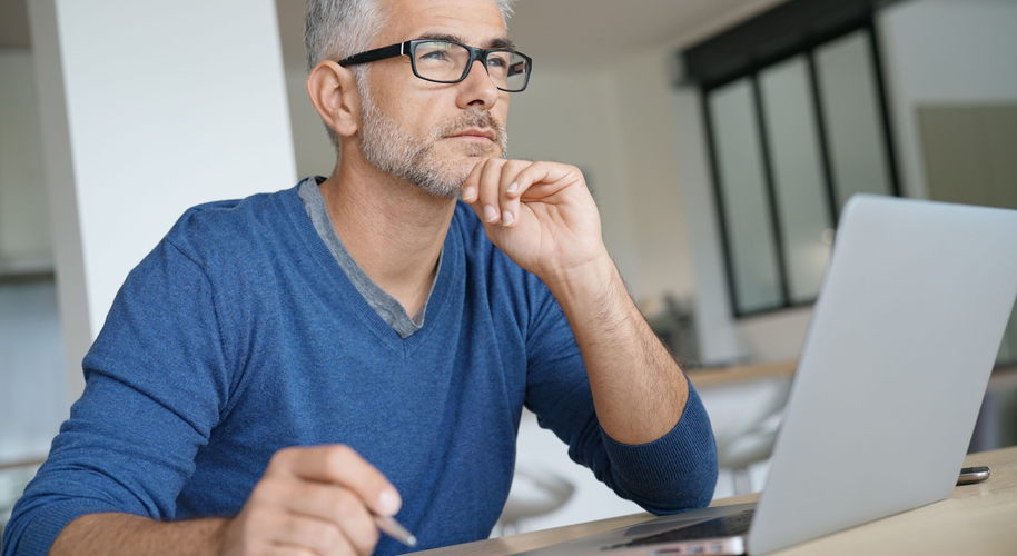 Grauhaariger Mann mit schwarzer Brille in blauem Pullover sitzt am Laptop und schaut in die Ferne