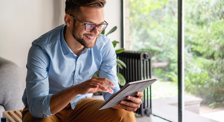 Mann in Jeanshemd und Chinohose sitzt in seinem Wohnzimmer und arbeitet am Tablet