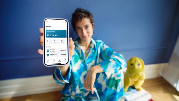 Frau in blauen Klamotten sitzt am Boden vor blauer Wand und hält ihr Smartphone mit der Consorsbank App in die Kamera