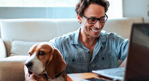 Mann mit Brille lacht und sitzt mit Hund vor dem Sofa und schaut auf Laptop