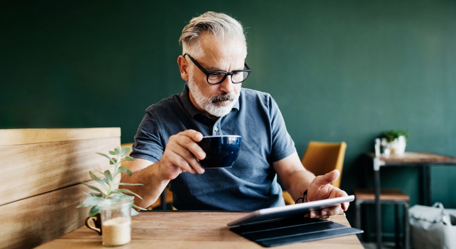 Mann mit grauen Haaren und schwarzer Brille schaut in sein Tablet und hält eine Tasse in der Hand